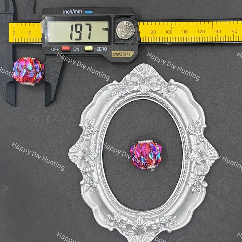 A faceted iridescent crystal bead shown with a silver ornate oval frame and a digital caliper on a dark gray background. The caliper reads 19.