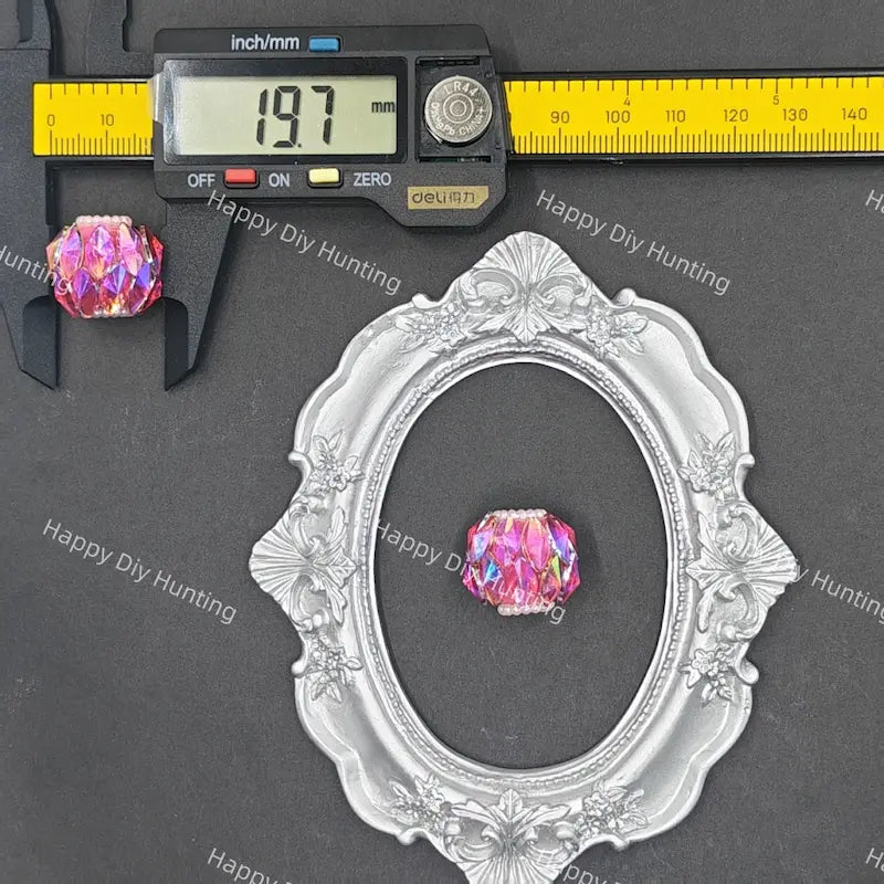 A pink iridescent faceted crystal bead shown with a silver ornate oval frame and a digital caliper on a dark gray background. The caliper reads 19.
