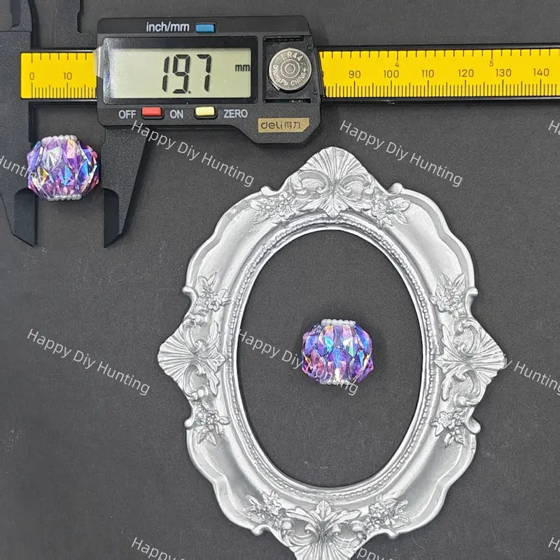 An oval silver-toned decorative frame displayed on a dark gray background, with two iridescent purple-blue beads shown beside it and a digital caliper above one bead to indicate size.