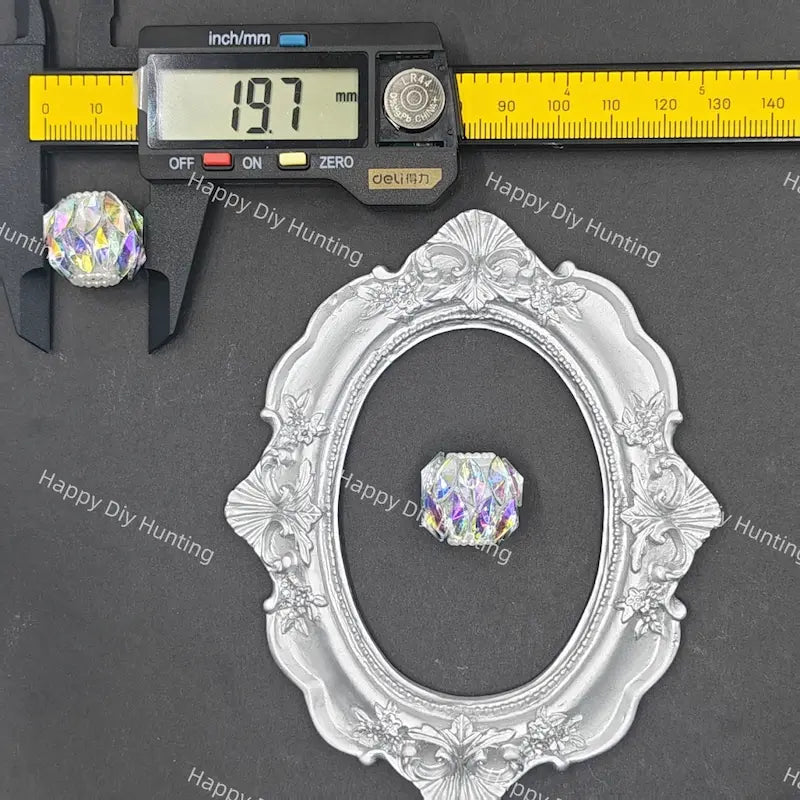 A silver ornate oval frame displayed on a dark gray background, with a small iridescent faceted crystal shown inside the frame and a digital caliper above it measuring 19.7 mm.