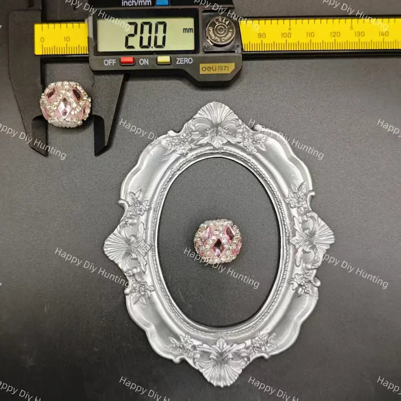 A pink rhinestone cabochon gemstone displayed on a black background, with a silver ornate oval frame and a digital caliper showing 20.0 mm measurements above it.
