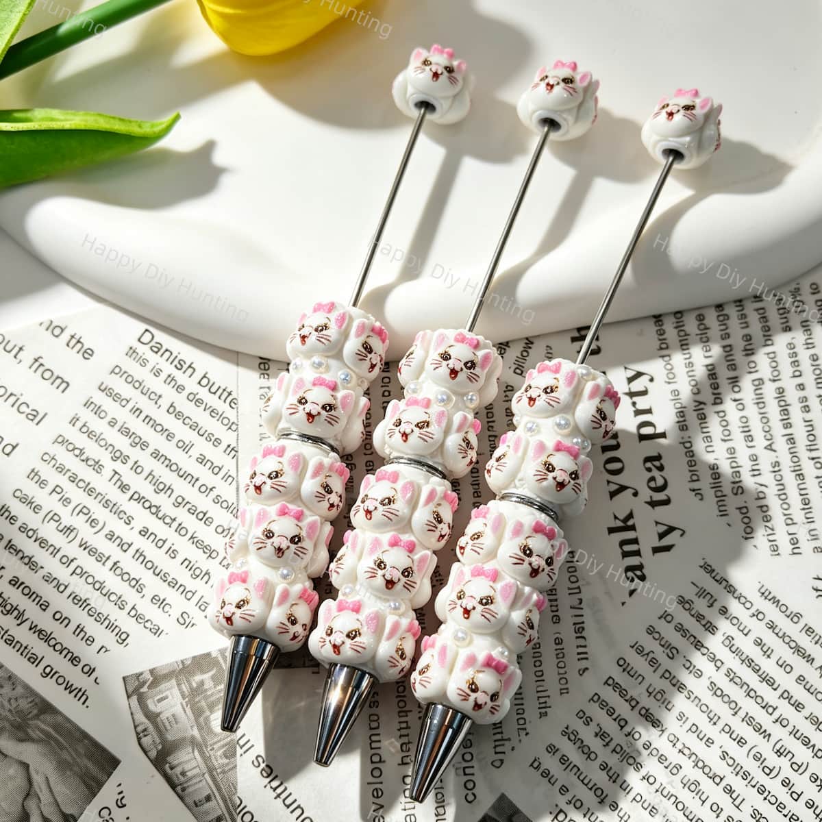 Three white cartoon bow cat character beadable pens displayed on a newspaper background. Each pen features white beads decorated with pink bow cat faces, faux pearls, and clear rhinestones, plus a silver metal tip. Perfect for DIY crafts, focal beads, and handmade gifts.
