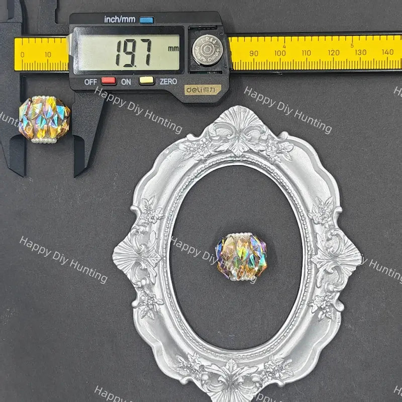A small oval faceted iridescent crystal bead is shown with a silver ornate frame and a digital caliper above it on a dark gray background. The caliper scale is marked in inches and millimeters, and the image includes repeated 'Happy DIY Hunting' watermarks.