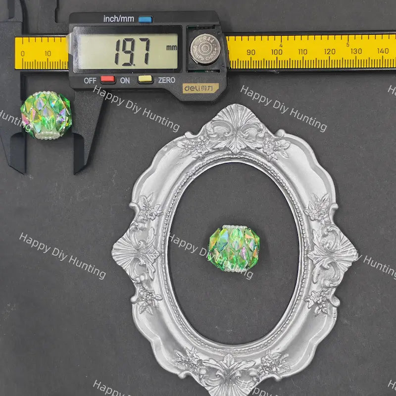 A green iridescent crystal bead shown with a silver ornate oval frame and a digital caliper on a dark gray background. The caliper displays 19.