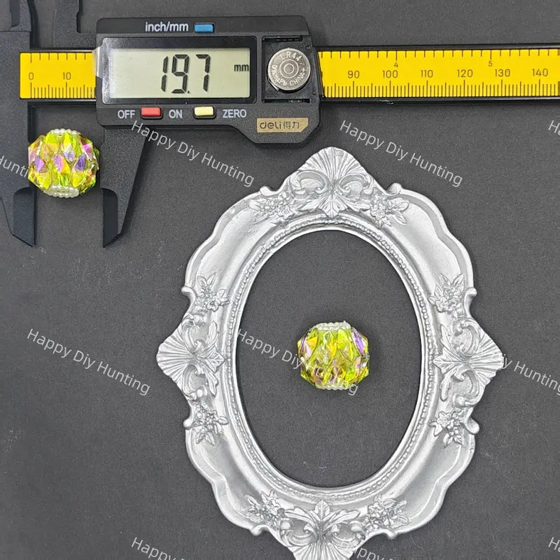 A small oval silver ornate frame or tray is shown on a dark gray background with a yellow digital caliper and a yellow measuring tape above it, alongside a small round iridescent yellow gemstone.