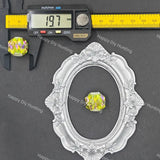 A small oval silver ornate frame or tray is shown on a dark gray background with a yellow digital caliper and a yellow measuring tape above it, alongside a small round iridescent yellow gemstone.
