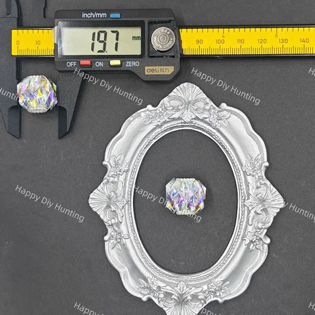 A silver ornate oval frame displayed on a dark gray background, with a small iridescent faceted crystal shown inside the frame and a digital caliper above it measuring 19.7 mm.