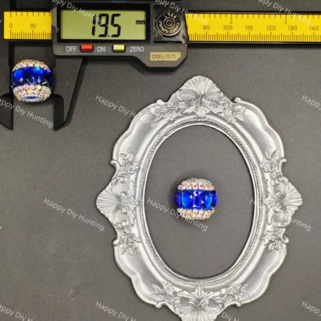 A blue and clear rhinestone ring displayed on an ornate silver oval frame, with a caliper at the top showing 19.5 mm measurements and a small ring on a black stand at the upper left.