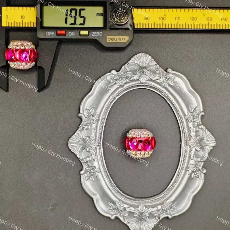 A silver ornate oval frame with a single pink and clear rhinestone bead at its center, shown next to a caliper measuring 19.5 mm.