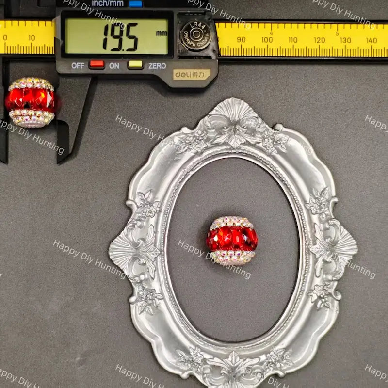 A red faceted crystal bead with a white rhinestone-encrusted border, displayed inside an ornate silver frame next to a digital caliper showing 19.5mm and a ruler showing 90mm.