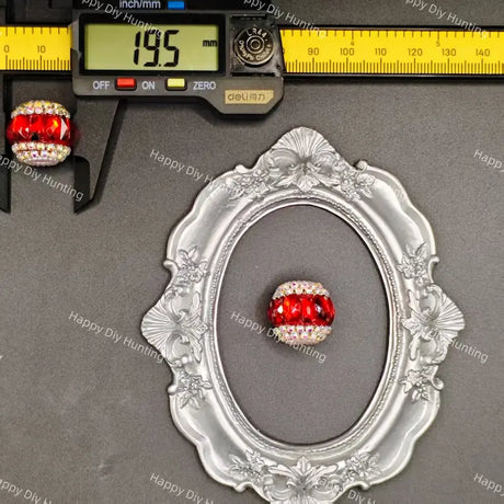 A red faceted crystal bead with a white rhinestone-encrusted border, displayed inside an ornate silver frame next to a digital caliper showing 19.5mm and a ruler showing 90mm.