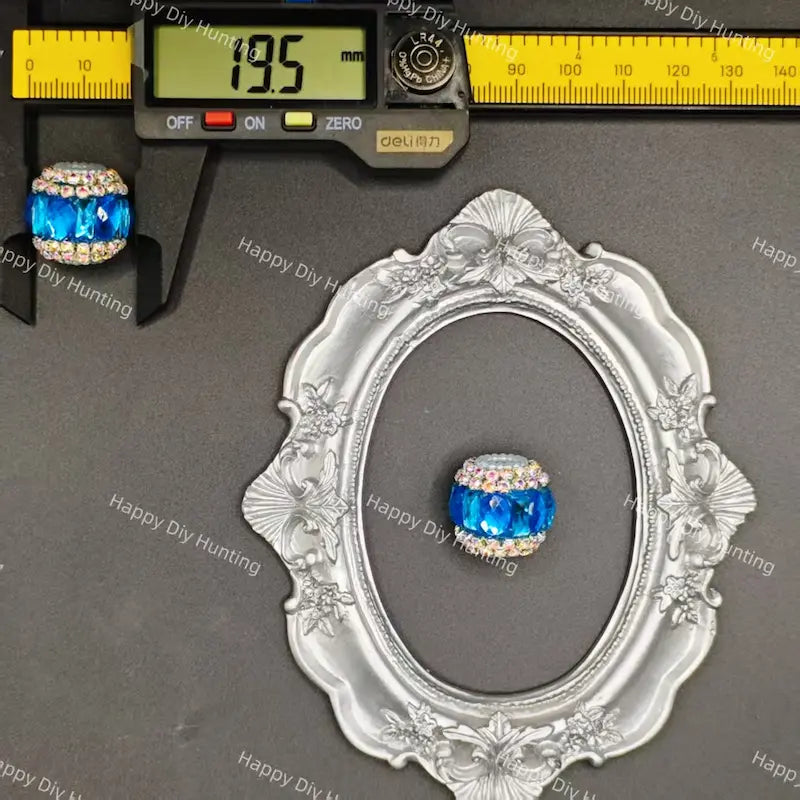 A blue faceted crystal bead with a rhinestone-encrusted top, shown alongside a digital caliper reading 19.5 mm and an ornate silver frame for scale, on a dark gray background with Happy DIY Hunting watermarks.