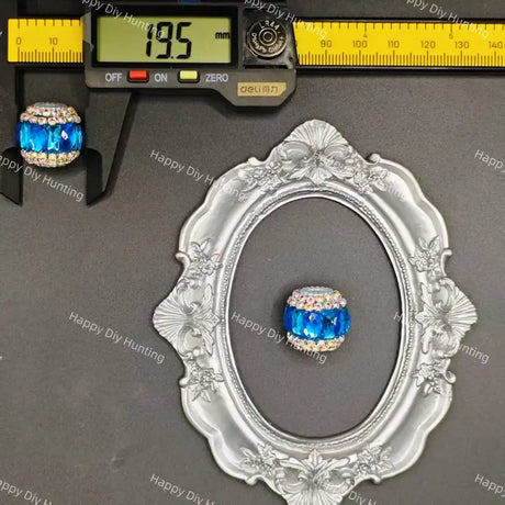 A blue faceted crystal bead with a rhinestone-encrusted top, shown alongside a digital caliper reading 19.5 mm and an ornate silver frame for scale, on a dark gray background with Happy DIY Hunting watermarks.