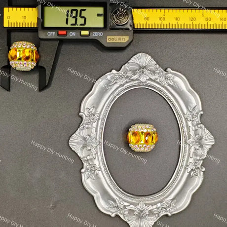 A rectangular yellow crystal bead with a silver-toned border and clear rhinestone accents, displayed on a gray background with a decorative silver frame and a caliper showing 19.5 mm measurement.
