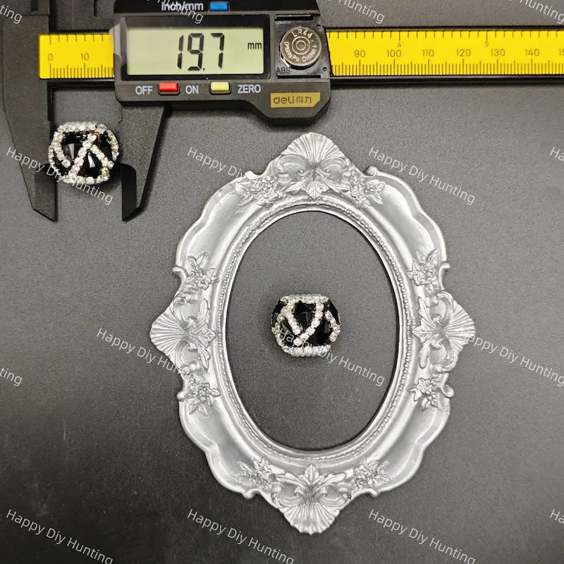A black and silver interlocking V ring displayed in an ornate silver oval frame, with a digital caliper showing 19.7 mm and a yellow ruler in the background.