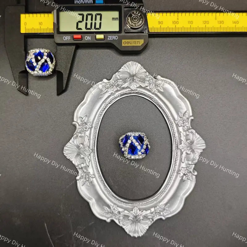 A ring with a blue and white geometric patterned center and clear crystal-like border, displayed on a dark surface with a decorative silver frame and a digital caliper above it for size reference. 