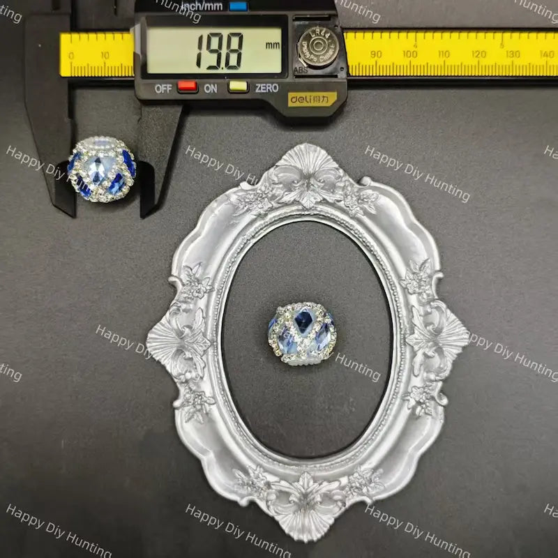 A small round blue and clear rhinestone bead or cabochon displayed on a dark gray surface, with a caliper above it showing 13.8 mm measurements and an ornate silver frame below for scale.