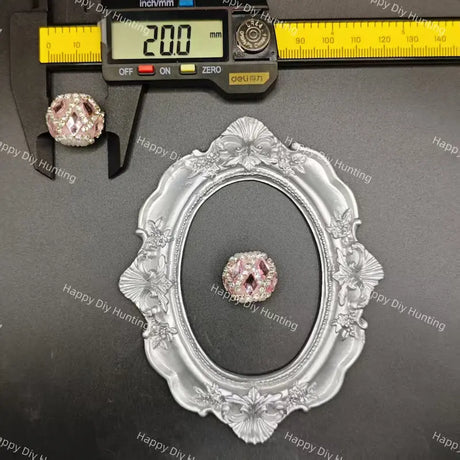 A pink rhinestone cabochon gemstone displayed on a black background, with a silver ornate oval frame and a digital caliper showing 20.0 mm measurements above it.