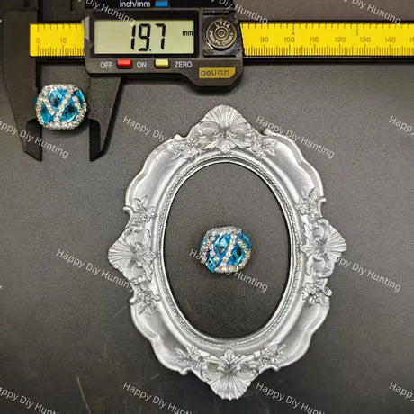 A ring with a blue and white patterned stone surrounded by clear crystals, displayed inside an ornate silver frame. A caliper is shown above it to indicate the ring's size, and the background is a dark gray surface with a repeating 'Happy DIY Hunting' watermark.