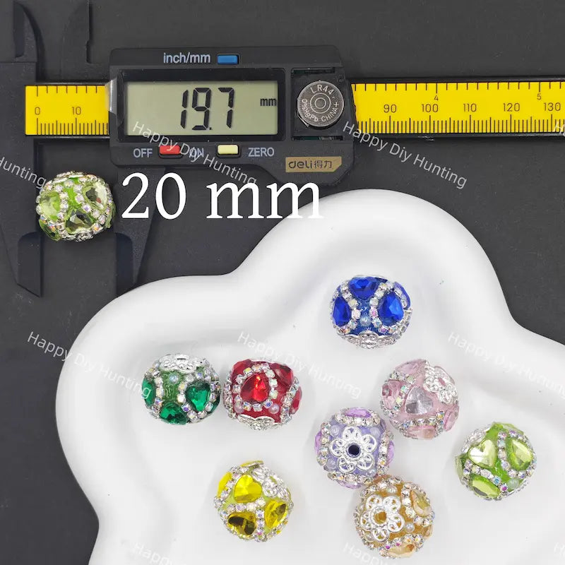 Seven colorful faceted crystal beads with silver-toned metal settings are displayed on a white cloud-shaped tray, alongside a digital caliper showing 20 mm measurement and a yellow scale ruler on a black background. 