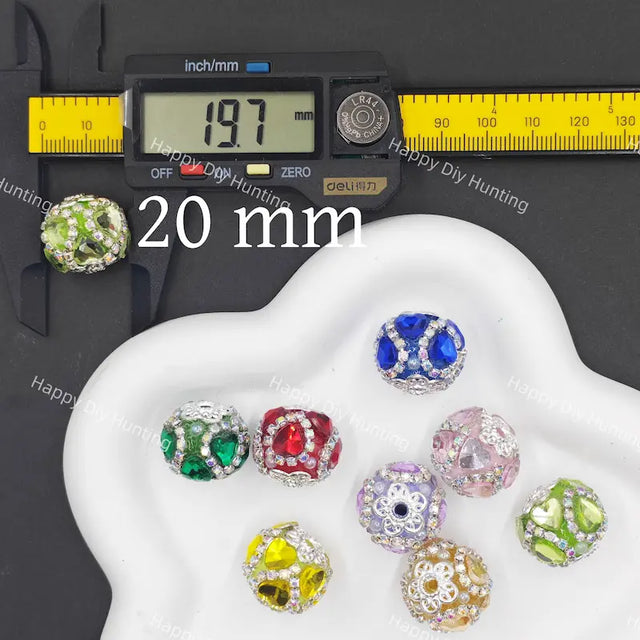 Seven colorful faceted crystal beads with silver-toned metal settings are displayed on a white cloud-shaped tray, alongside a digital caliper showing 20 mm measurement and a yellow scale ruler on a black background. 