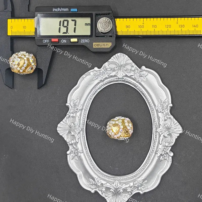 A small oval silver frame displayed on a dark gray background, with a yellow caliper above it showing 19.7 mm measurements and a gold-and-white beaded ornament placed inside the frame for scale.
