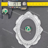 A small oval silver frame with ornate floral detailing is shown on a dark gray background, with a matching green and white gemstone placed in the center.