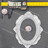 A silver oval decorative frame with ornate floral detailing, shown on a dark gray background with a digital caliper measuring 197 mm and a small pink faceted gemstone placed inside the frame for scale.