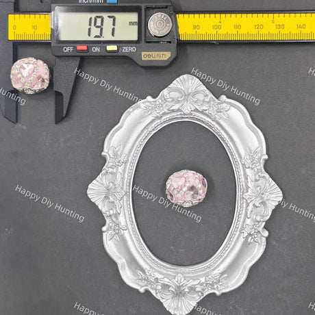 A silver oval decorative frame with ornate floral detailing, shown on a dark gray background with a digital caliper measuring 197 mm and a small pink faceted gemstone placed inside the frame for scale.