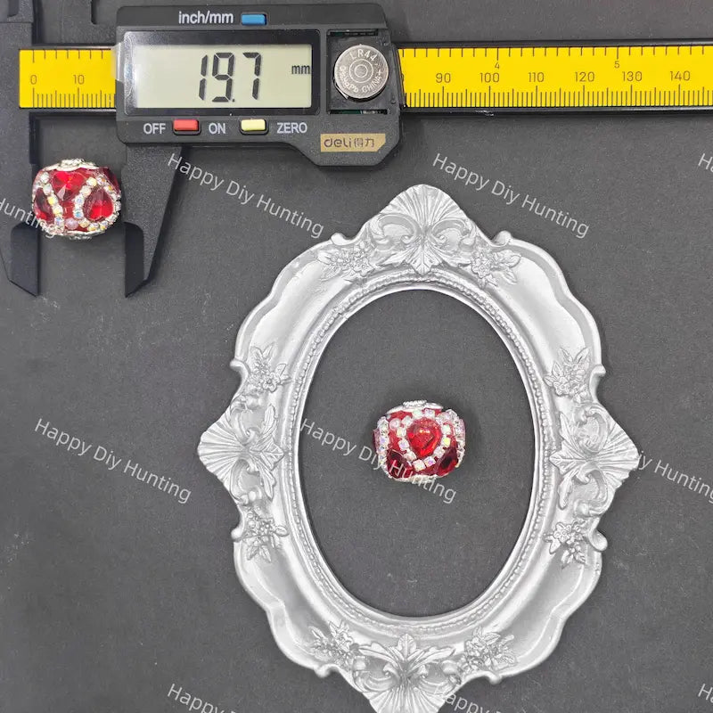 A small red and white beaded jewelry charm is shown on a dark gray background with a silver ornate oval frame, a digital caliper reading 19.7 mm, and a yellow inch/mm ruler.