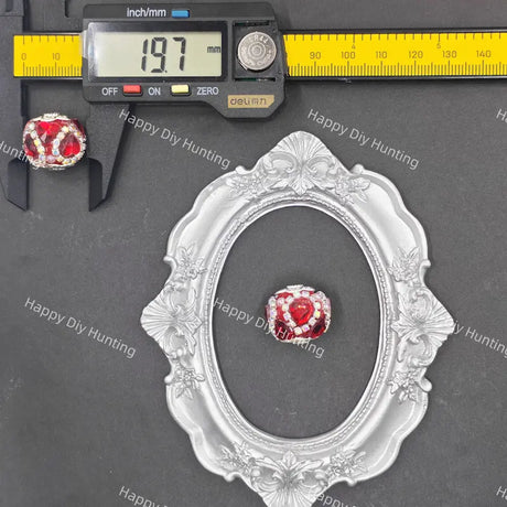 A small red and white beaded jewelry charm is shown on a dark gray background with a silver ornate oval frame, a digital caliper reading 19.7 mm, and a yellow inch/mm ruler.