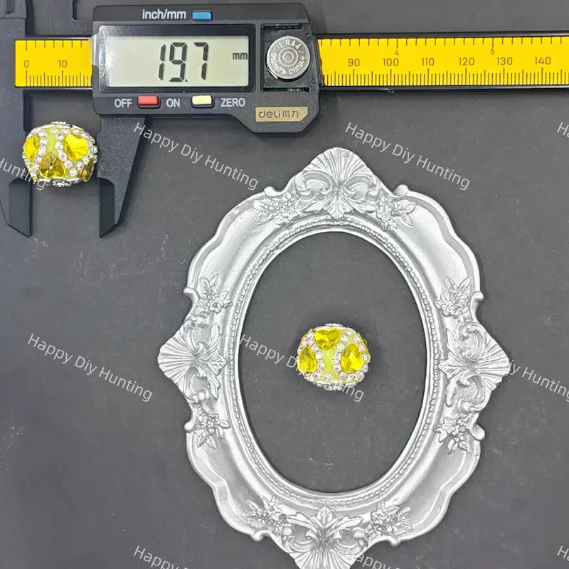 A small oval ring with a yellow center and clear surrounding stones is shown on a dark gray surface, alongside a silver ornate frame, a digital caliper reading 19.7 mm, and repeated Happy DIY Hunting watermarks.