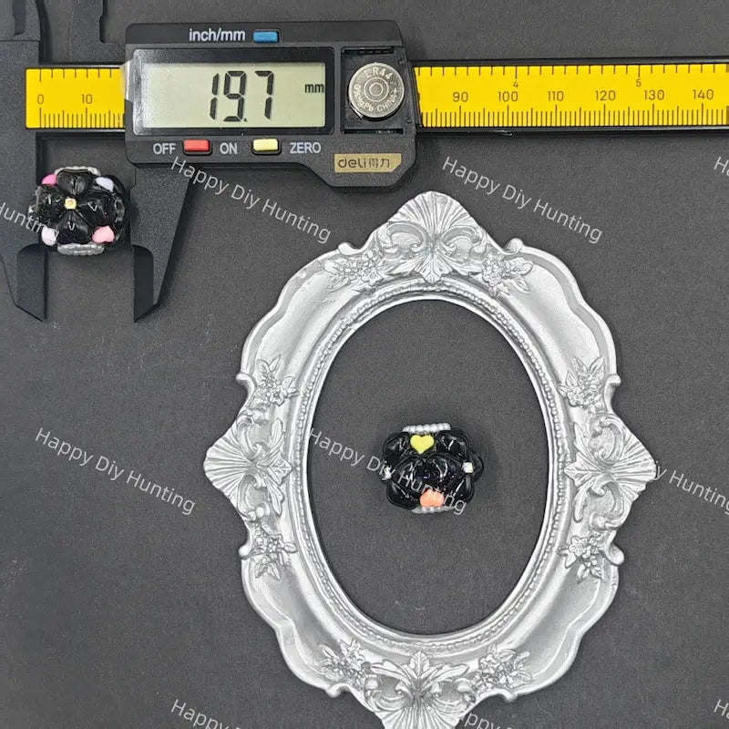 A small black and white cat figurine with yellow and pink details, shown next to a digital caliper reading 19.7mm and an ornate silver oval frame for scale, on a dark gray background with Happy Diy Hunting watermarks.