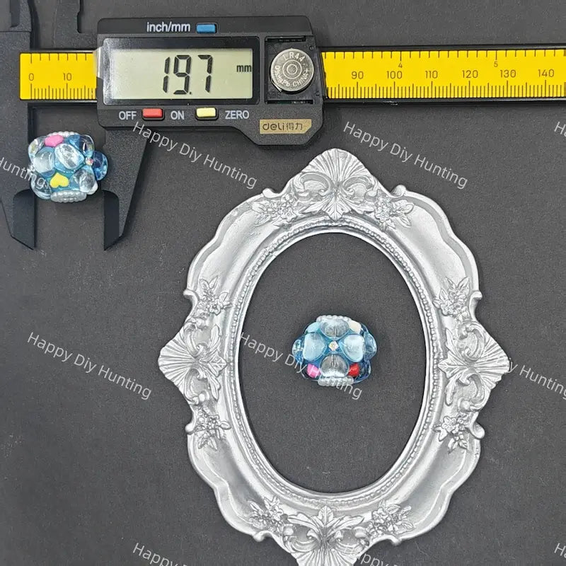 A decorative silver oval frame displayed on a dark gray background with a yellow caliper and digital ruler for scale, alongside a small blue and pink skull-shaped ornament.