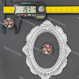 A small decorative oval frame and a matching oval-shaped jewelry piece are shown on a dark gray surface with a digital caliper and yellow ruler for scale. The jewelry appears to be a cluster of brown stones with light blue and pink accents.