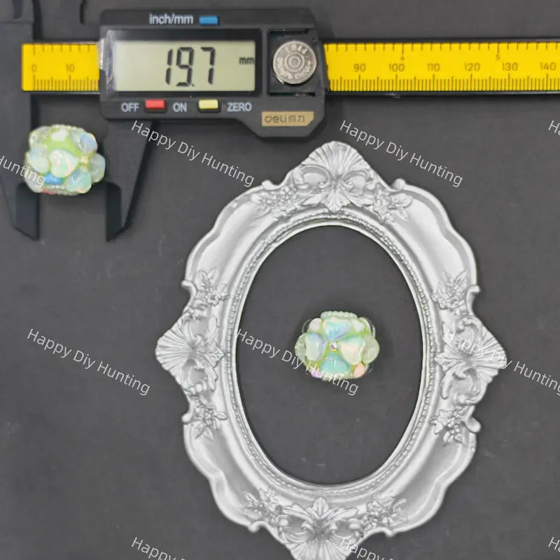 A small iridescent opal-like stone or bead shown with a silver ornate frame, a digital caliper reading 19.7 mm, and a yellow ruler on a dark gray background.