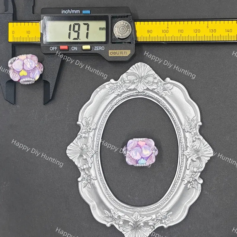 A small iridescent lavender and pink flower-shaped bead or charm displayed on a dark gray background, with a silver ornate oval frame, a yellow caliper, and a digital ruler above it. Repeated 'Happy Diy Hunting' watermarks are visible across the photo.