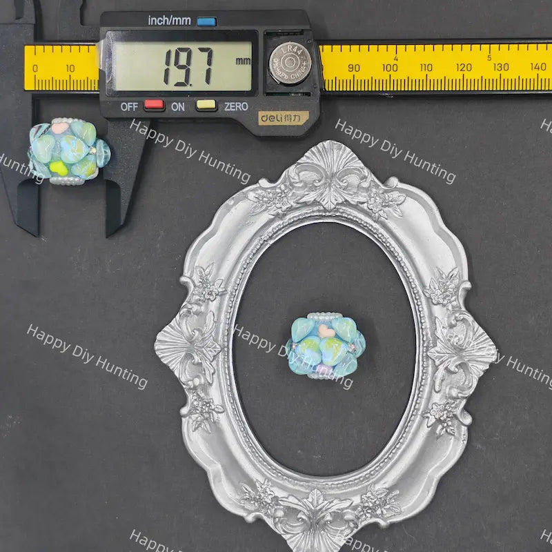 A decorative silver oval frame displayed on a dark gray surface, with a small blue-green flower-shaped bead placed inside the center. A black caliper and a yellow measuring ruler are shown above to indicate scale, and the word 'deublin' is visible on the measuring tool.