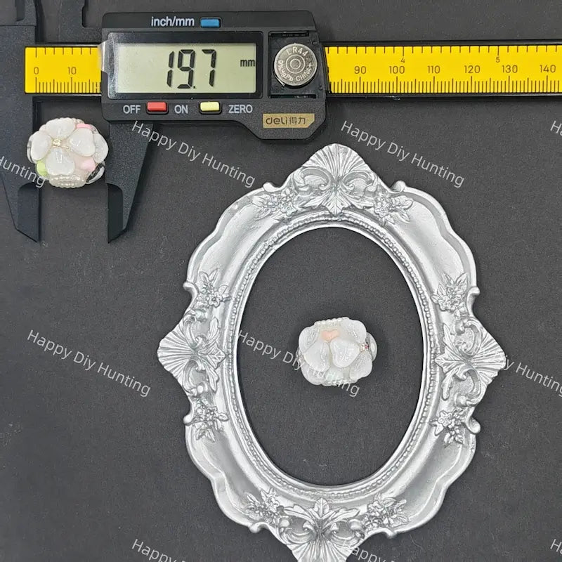 An ornate silver oval picture frame with floral scrollwork, shown on a dark gray background with a digital caliper measuring 19.7 mm and a small white flower accent.
