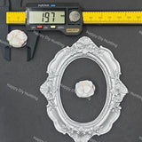 An ornate silver oval picture frame with floral scrollwork, shown on a dark gray background with a digital caliper measuring 19.7 mm and a small white flower accent.