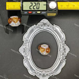 A small ornate silver oval frame with floral detailing is shown on a dark gray background, with a jeweled bead or charm placed inside the center