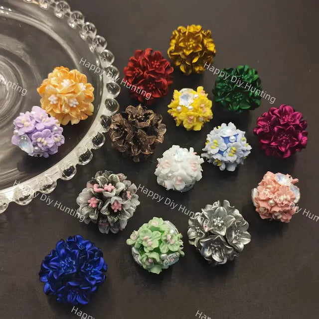 A collection of colorful, flower-shaped jewelry findings or beads arranged on a dark surface next to a clear glass dish. The set includes various colors such as blue, red, green, yellow, purple, pink, white, and silver.