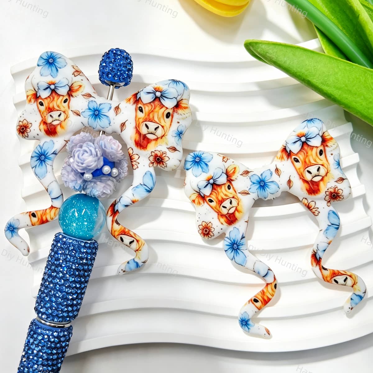 Blue highland cow floral print silicone bow beads displayed with a blue rhinestone beadable pen. Featuring an adorable highland cow with a bow and blue flowers. Soft, durable, and perfect for beadable pens, keychains, and handmade crafts