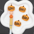 Four orange pumpkin-shaped charms with 'BOO!' written on them in black, arranged on a white cloud-shaped palette next to a paintbrush.