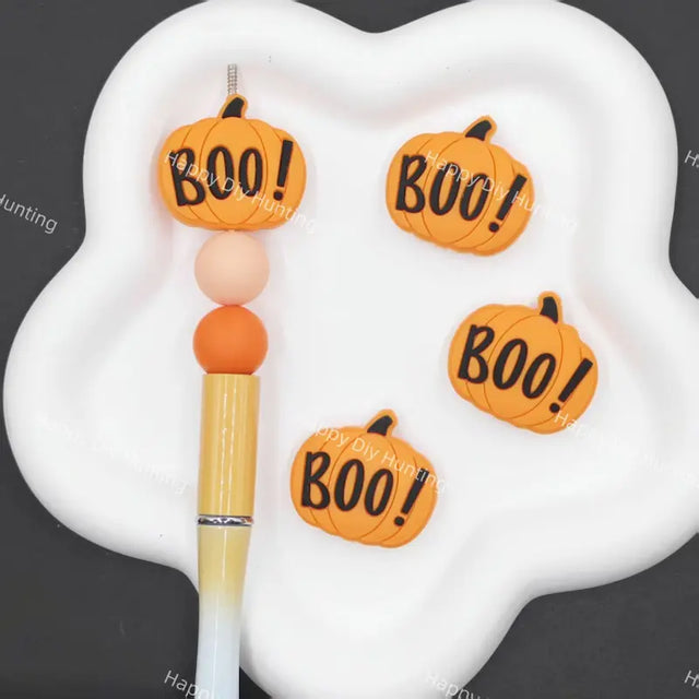 Four orange pumpkin-shaped charms with 'BOO!' written on them in black, arranged on a white cloud-shaped palette next to a paintbrush.