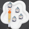 Three cartoon ghost charms with blue hats and yellow glasses are displayed on a white cloud-shaped tray next to a pen, on a dark gray background.