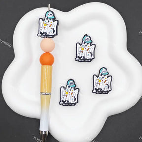 Three cartoon ghost charms with blue hats and yellow glasses are displayed on a white cloud-shaped tray next to a pen, on a dark gray background.