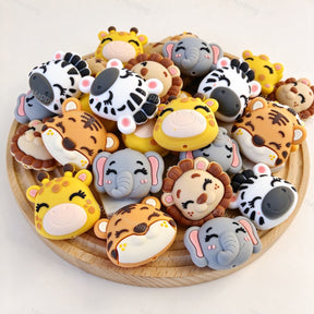 A mixed pile of cute safari animal silicone focal beads on a wooden tray. Designs include tiger, giraffe, lion, zebra, and elephant, all with closed eyes and sweet smiles. Soft, durable, and perfect for beadable pens, keychains, and handmade crafts.