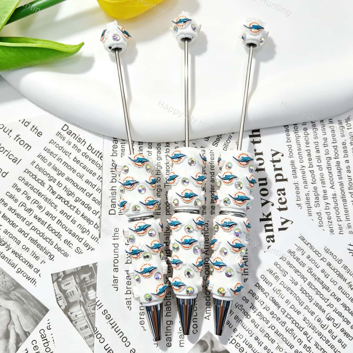 Three dolphin sports team logo print sports beadable pens displayed on a newspaper background. Each pen features white beads decorated with teal dolphin and orange sun team logos, plus iridescent rhinestones and silver metal tips. Perfect for sports fans, DIY crafts, focal beads, and handmade gifts.