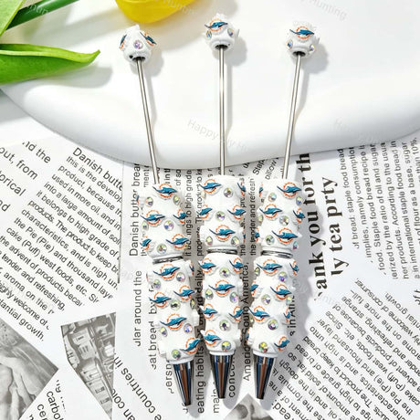 Three dolphin sports team logo print sports beadable pens displayed on a newspaper background. Each pen features white beads decorated with teal dolphin and orange sun team logos, plus iridescent rhinestones and silver metal tips. Perfect for sports fans, DIY crafts, focal beads, and handmade gifts.